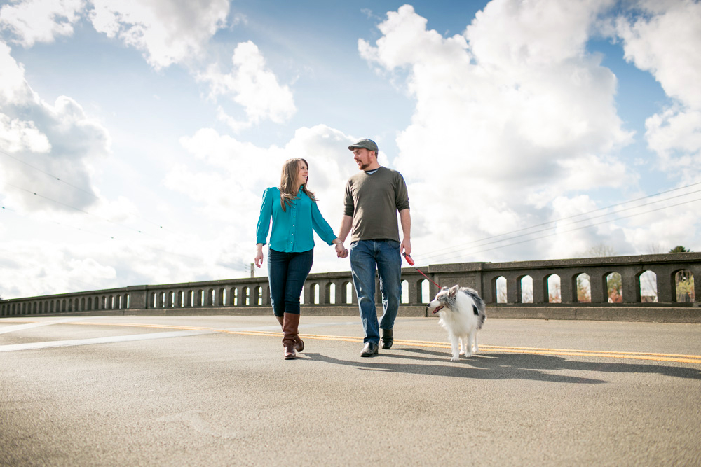 A man and woman holding hands walk a dog together on a sunny day along a road with a concrete barrier and a cloudy sky in the background. Jessica Hill Photography - Wedding Photographer - Oregon