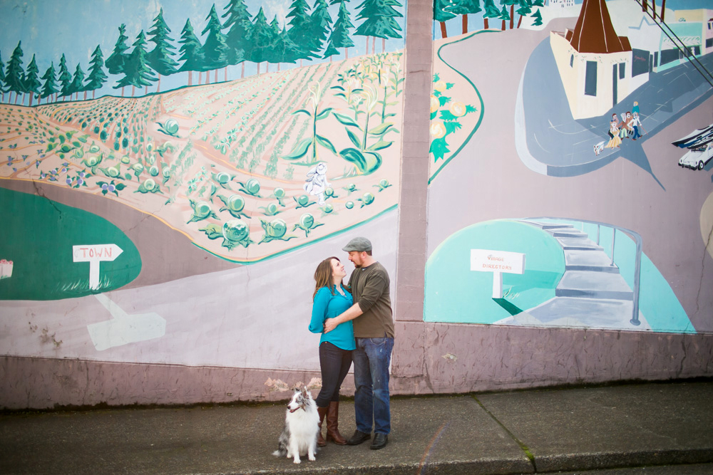 A couple stands close together on a sidewalk with a dog, in front of a large, colorful mural depicting a town and fields. Jessica Hill Photography - Wedding Photographer - Oregon