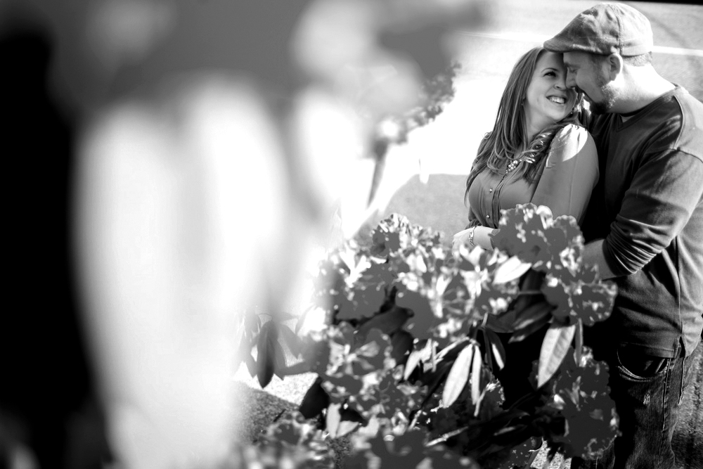 A man and woman stand close together outdoors, smiling and embracing beside leafy plants. The image is in black and white. Jessica Hill Photography - Wedding Photographer - Oregon
