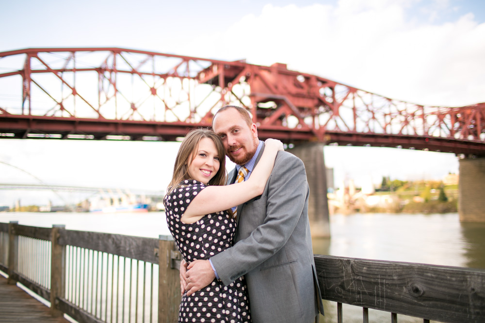 A woman and man embrace and smile at the camera while standing on a wooden walkway by a river, with a large red metal bridge in the background. Jessica Hill Photography - Wedding Photographer - Oregon
