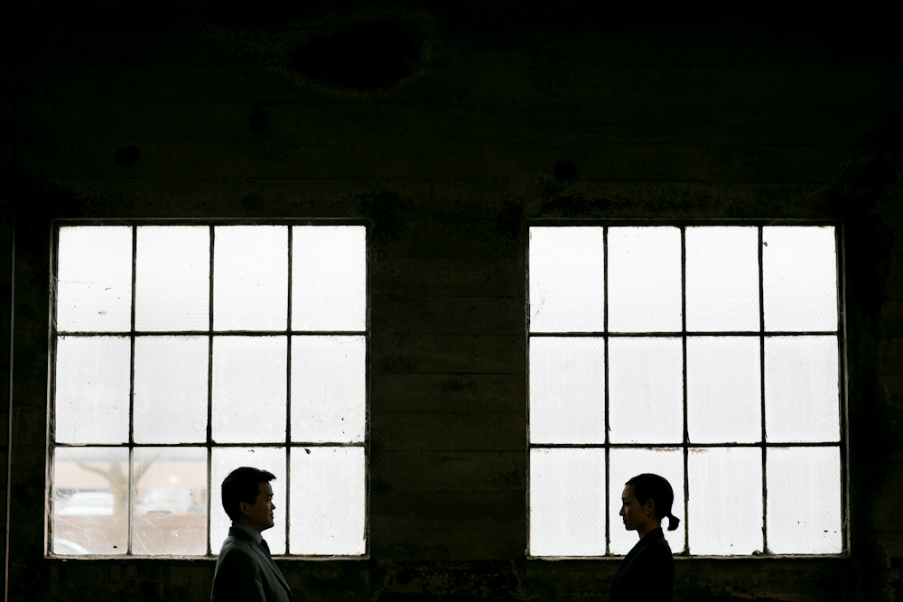 Two people in profile face each other, silhouetted against two large industrial windows in a dimly lit room. Jessica Hill Photography - Wedding Photographer - Oregon