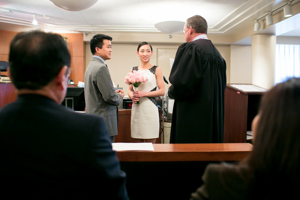 A couple stands before a judge holding hands during a wedding ceremony in a courtroom, with two people seated in the foreground. Jessica Hill Photography - Wedding Photographer - Oregon