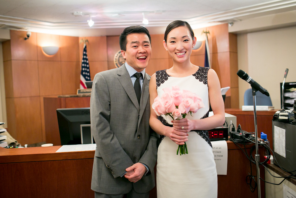 A smiling couple poses together in a courtroom; the woman holds a bouquet of pink roses and wears a white dress, while the man is dressed in a gray suit. Jessica Hill Photography - Wedding Photographer - Oregon