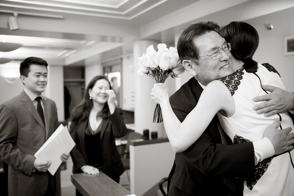 A woman hugs an older man holding a bouquet of flowers while two people in business attire smile and observe in an indoor setting. Jessica Hill Photography - Wedding Photographer - Oregon