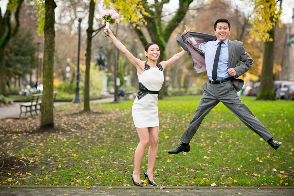 A woman in a white dress and a man in a suit jump and smile in a park, with green grass and autumn trees in the background. Jessica Hill Photography - Wedding Photographer - Oregon