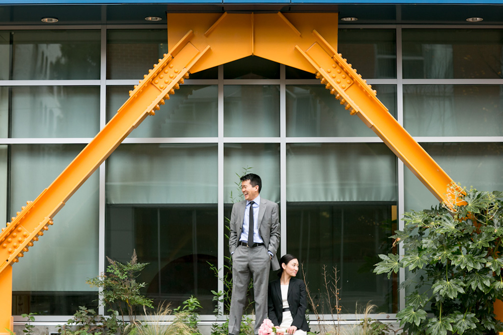 Two people in business attire stand near an orange structural beam in front of a modern glass building with plants nearby. Jessica Hill Photography - Wedding Photographer - Oregon