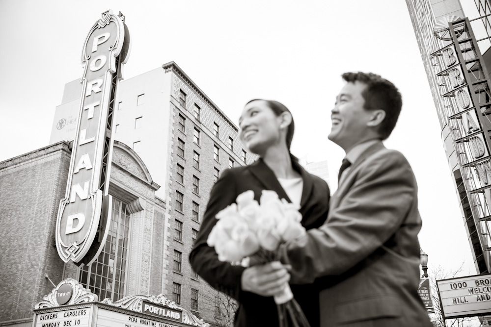 A smiling couple hugs at a lively indoor event, surrounded by well-dressed guests, some with raised arms in celebration. Jessica Hill Photography - Wedding Photographer - Oregon