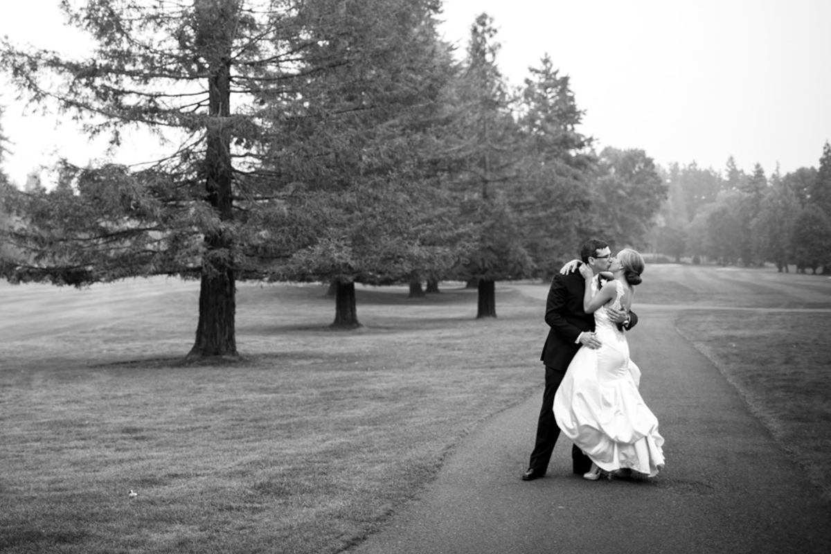 A bride and groom embrace and kiss on a paved path surrounded by grass and tall trees in a park. The bride wears a white dress and the groom wears a suit. The photo is black and white. Jessica Hill Photography - Wedding Photographer - Oregon
