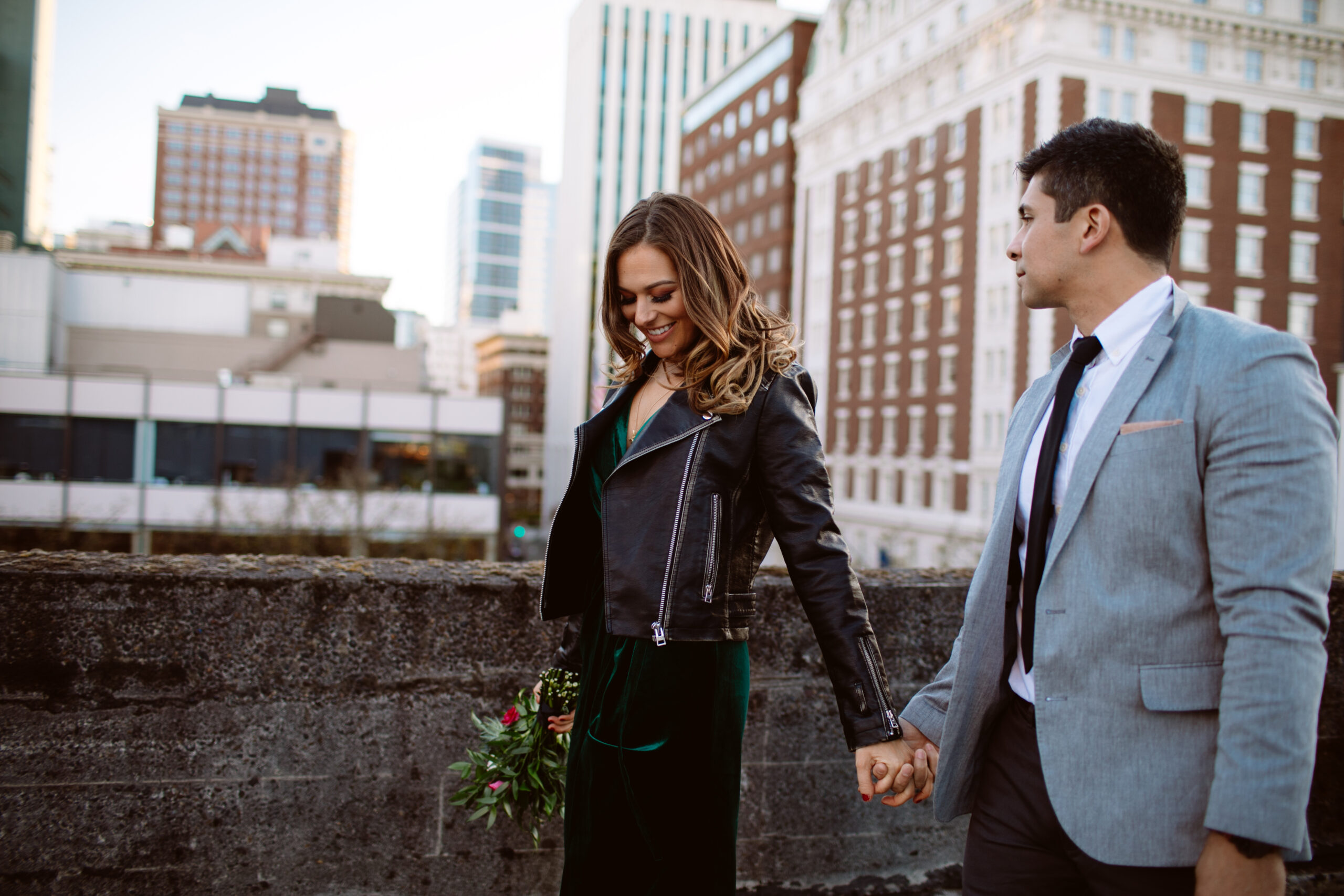 A woman holding a bouquet and a man in a suit hold hands while walking on a rooftop in a city, with tall buildings in the background. Jessica Hill Photography
