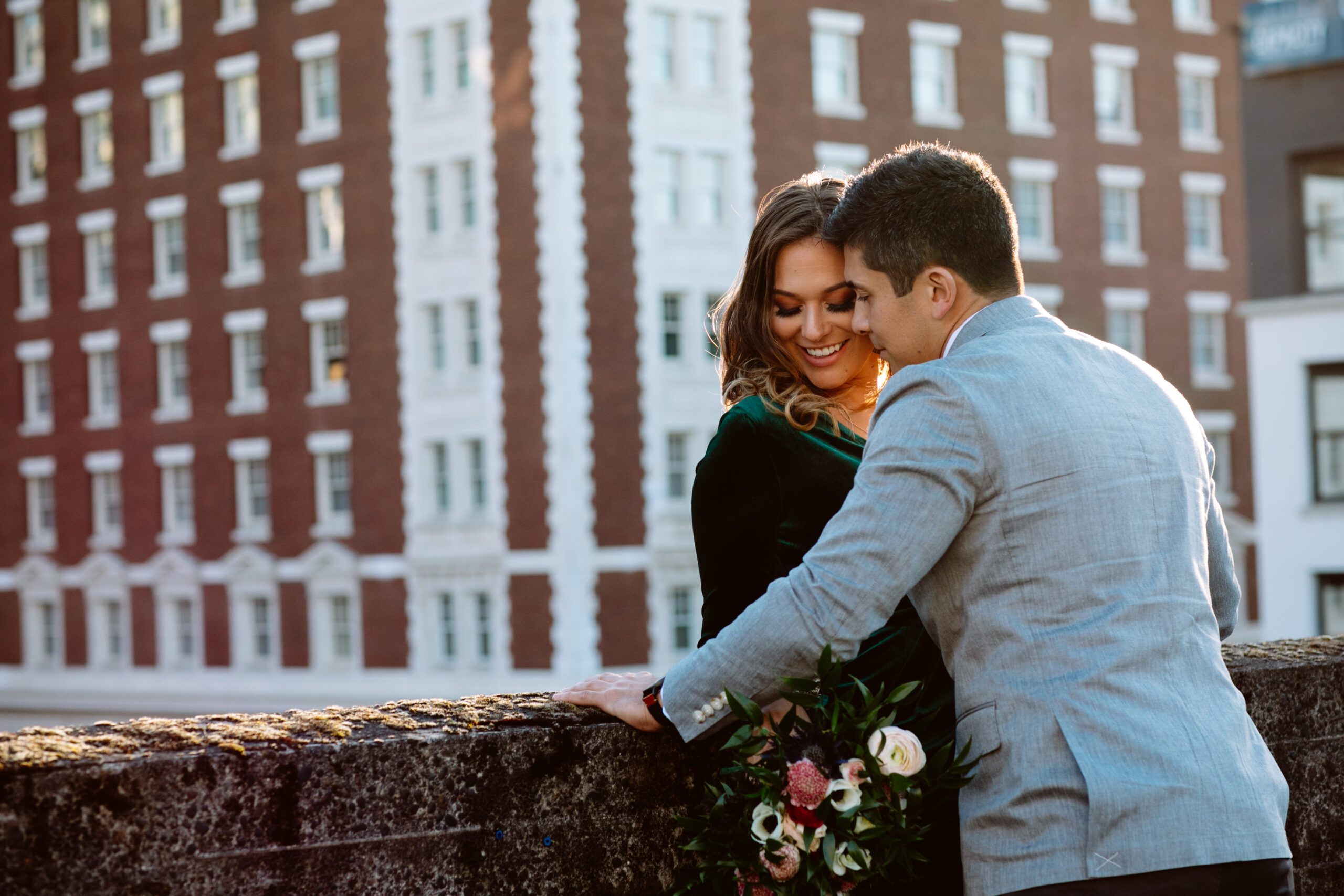 A couple stands close together by a stone wall, holding a bouquet, with a tall brick building in the background. Jessica Hill Photography