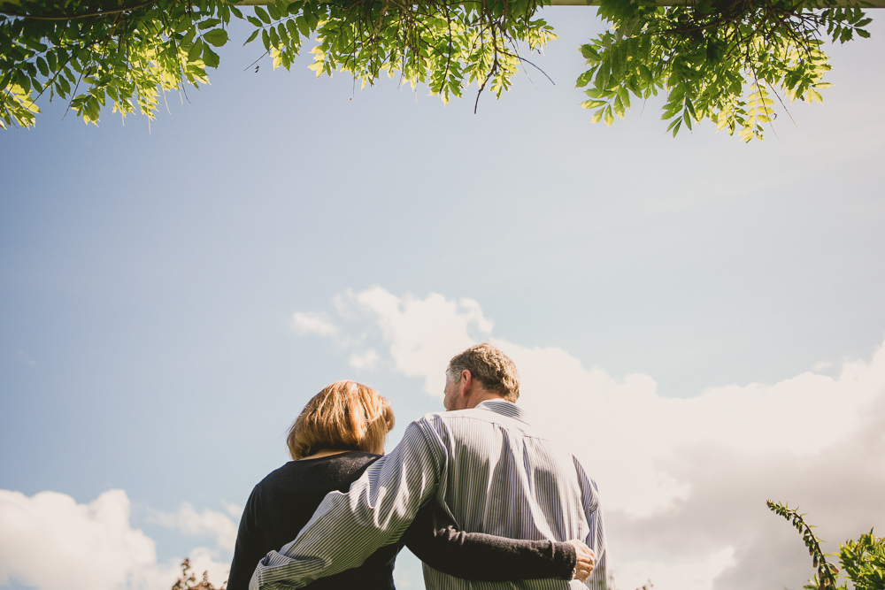 Two people stand closely together outdoors, viewed from behind, with trees and blue sky with clouds in the background. Jessica Hill Photography - Wedding Photographer - Oregon