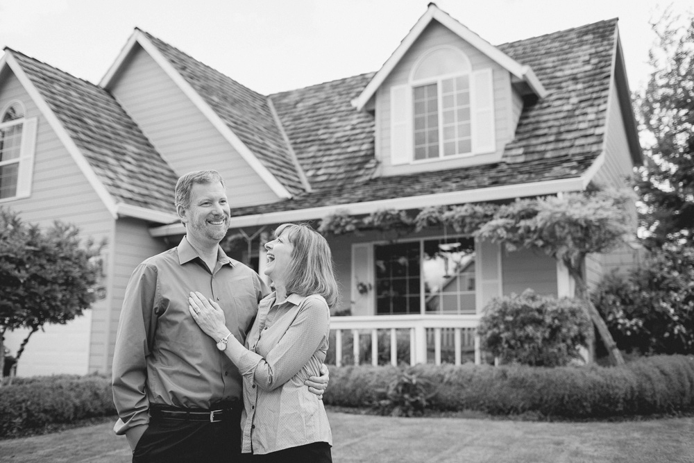 A man and woman stand smiling together in front of a house with a gabled roof and large windows; the image is in black and white. Jessica Hill Photography - Wedding Photographer - Oregon