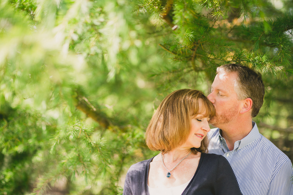A man kisses a woman's head while standing together outdoors in front of green pine branches. Both appear relaxed and content. Jessica Hill Photography - Wedding Photographer - Oregon