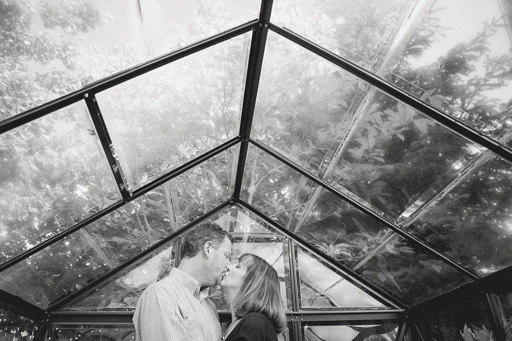 A couple stands closely facing each other, about to kiss, inside a glass greenhouse with trees visible through the roof above them. Jessica Hill Photography - Wedding Photographer - Oregon