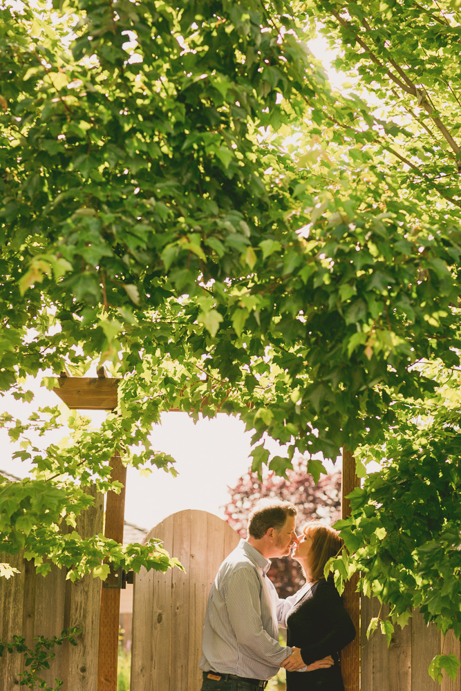 A man and woman stand close together under leafy trees by a wooden fence on a sunny day. Jessica Hill Photography - Wedding Photographer - Oregon