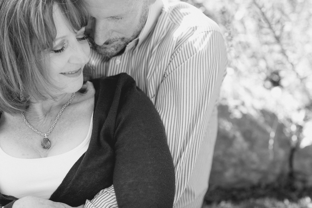 A man embraces a woman from behind as they both smile, sitting outdoors in natural light. The image is in black and white. Jessica Hill Photography - Wedding Photographer - Oregon