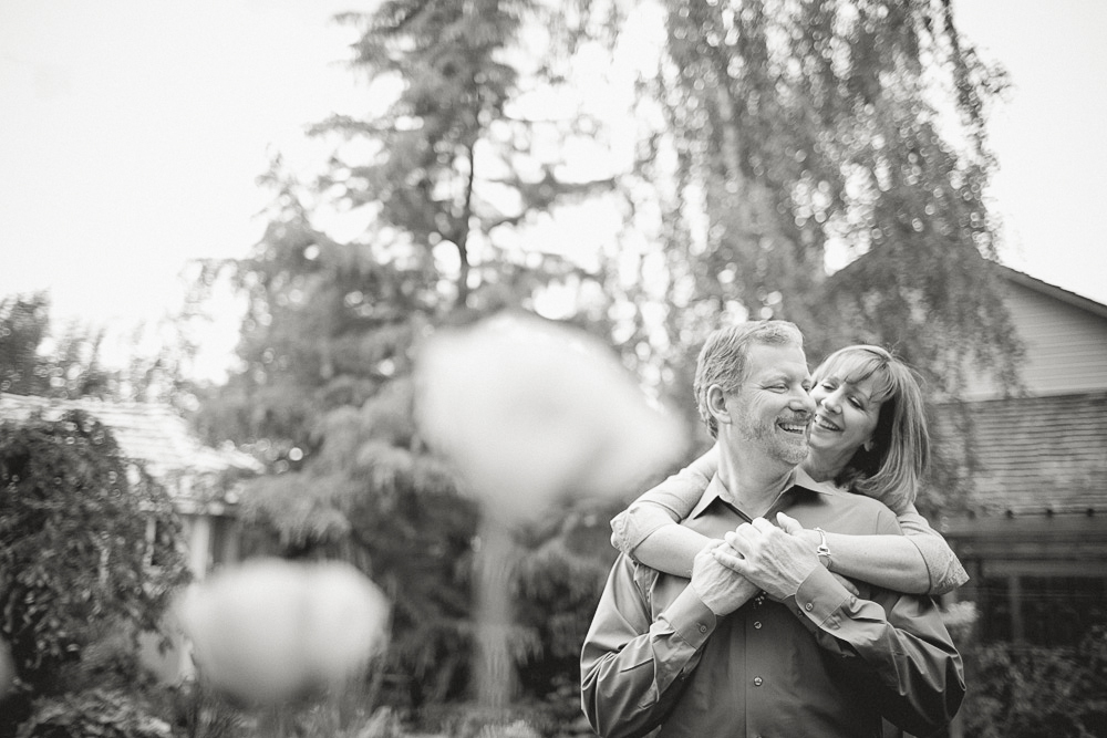 A woman embraces a smiling man from behind in an outdoor garden setting with trees and a house visible in the background. Jessica Hill Photography - Wedding Photographer - Oregon