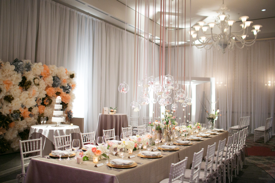 Elegant wedding reception with a long dining table, floral centerpieces, hanging glass ornaments, white chairs, and a flower wall. A tiered wedding cake stands on a side table. Jessica Hill Photography - Wedding Photographer - Oregon
