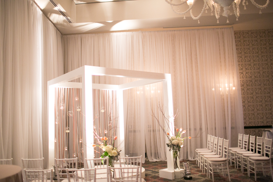A modern indoor wedding setup featuring a white chuppah with floral arrangements, surrounded by rows of white chairs and soft draped curtains. Jessica Hill Photography - Wedding Photographer - Oregon