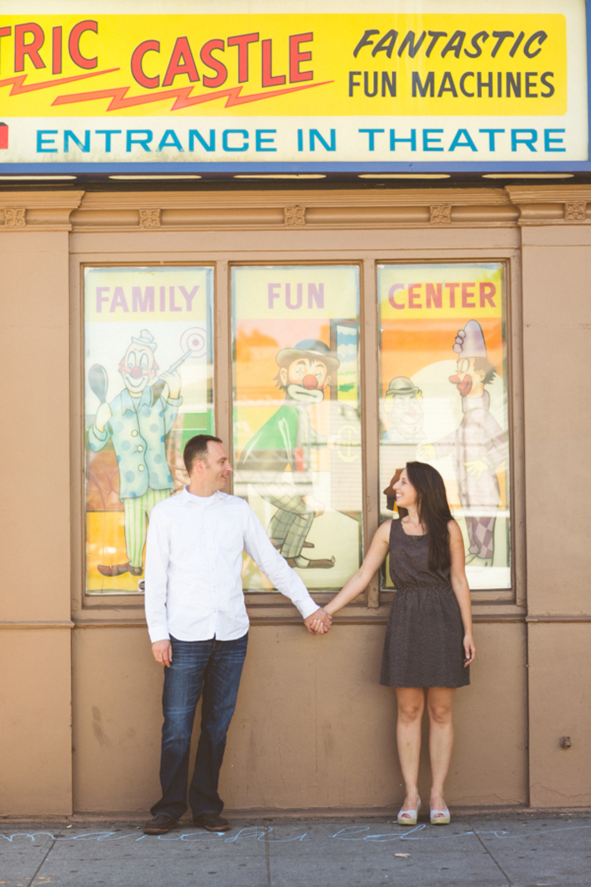 A couple sits at a table in a cafe, smiling and leaning close to each other while holding hands. The scene is viewed through a window reflecting the surroundings. Jessica Hill Photography