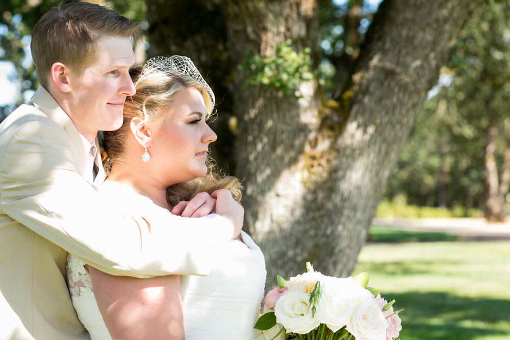 A bride and groom stand outdoors in sunlight, with the groom embracing the bride from behind. She holds a bouquet of white and pale pink flowers. Trees are visible in the background. Jessica Hill Photography - Wedding Photographer - Oregon