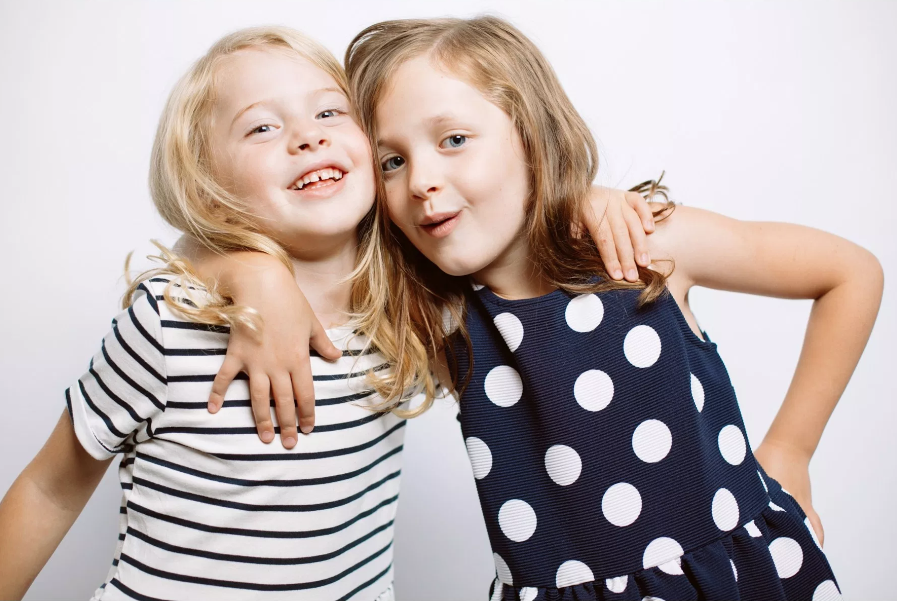 Two young girls stand close together, smiling at the camera. One girl wears a striped shirt, and the other wears a polka dot dress. Jessica Hill Photography