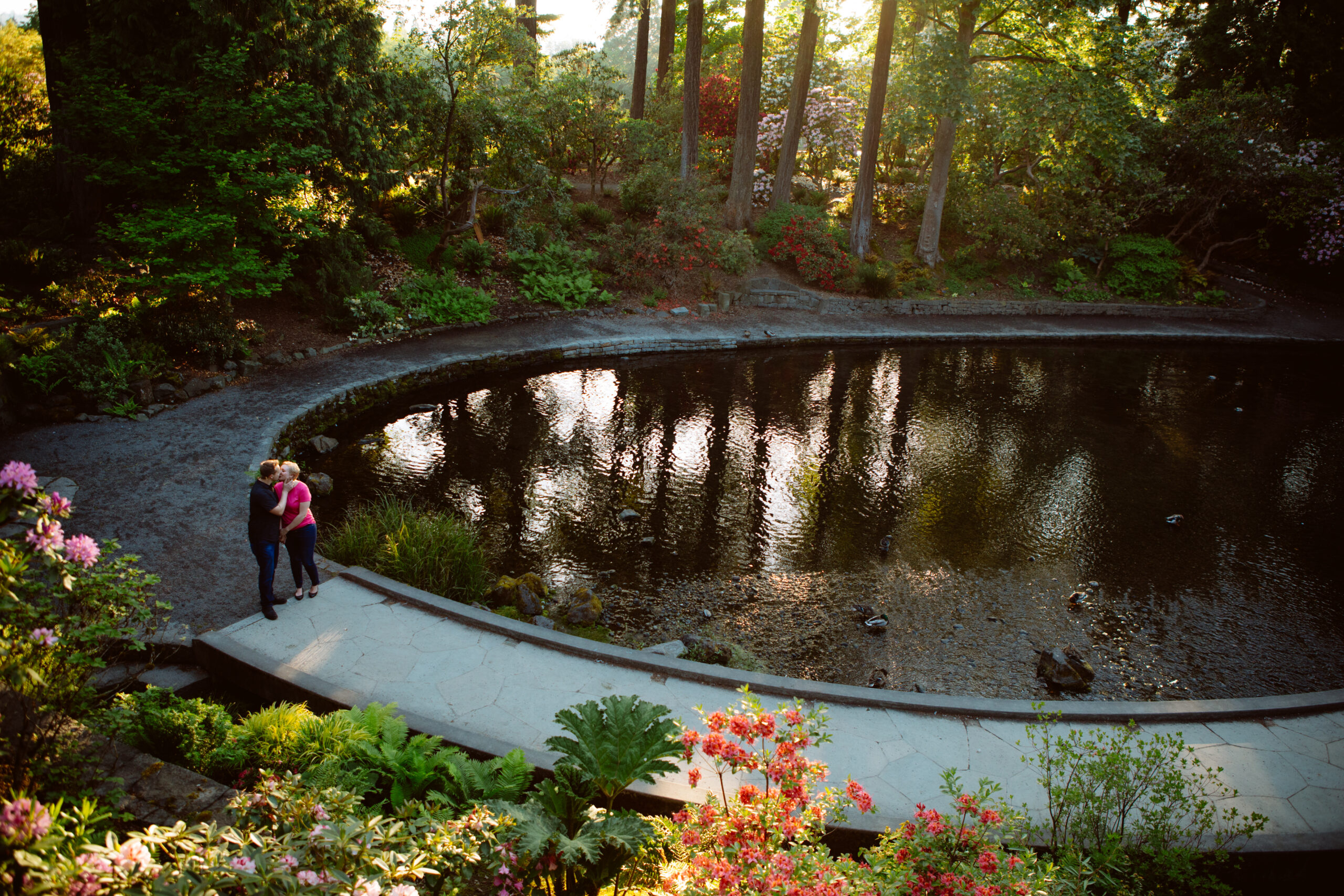 Two people stand on a curved path beside a reflective pond surrounded by lush greenery and tall trees in a sunlit garden. Jessica Hill Photography