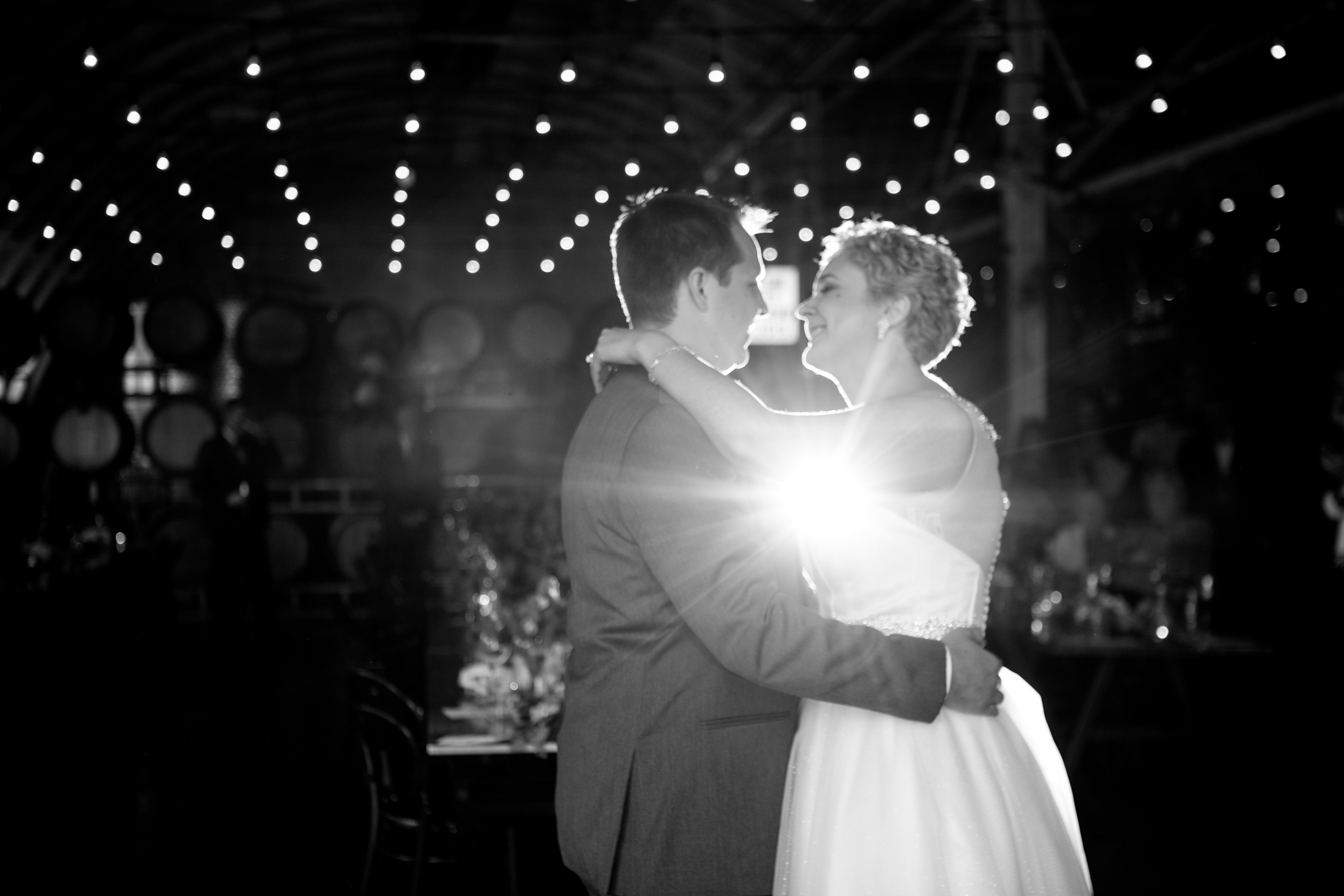 A bride and groom share a dance under string lights, with a bright light shining between them in a dimly lit venue. Jessica Hill Photography