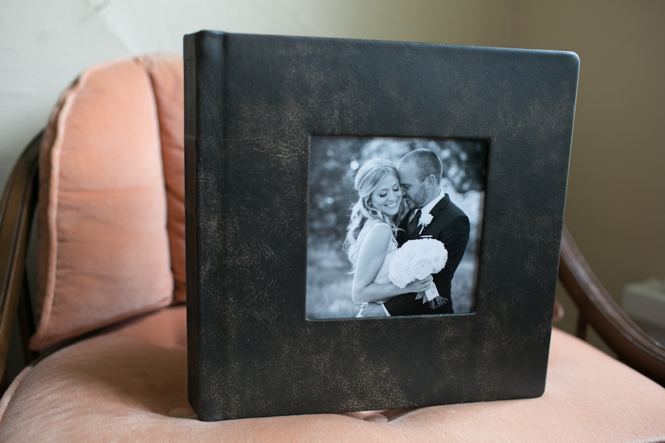 A black leather photo album with a cover image of a bride and groom embracing, placed on a pink upholstered chair. Jessica Hill Photography - Wedding Photographer - Oregon