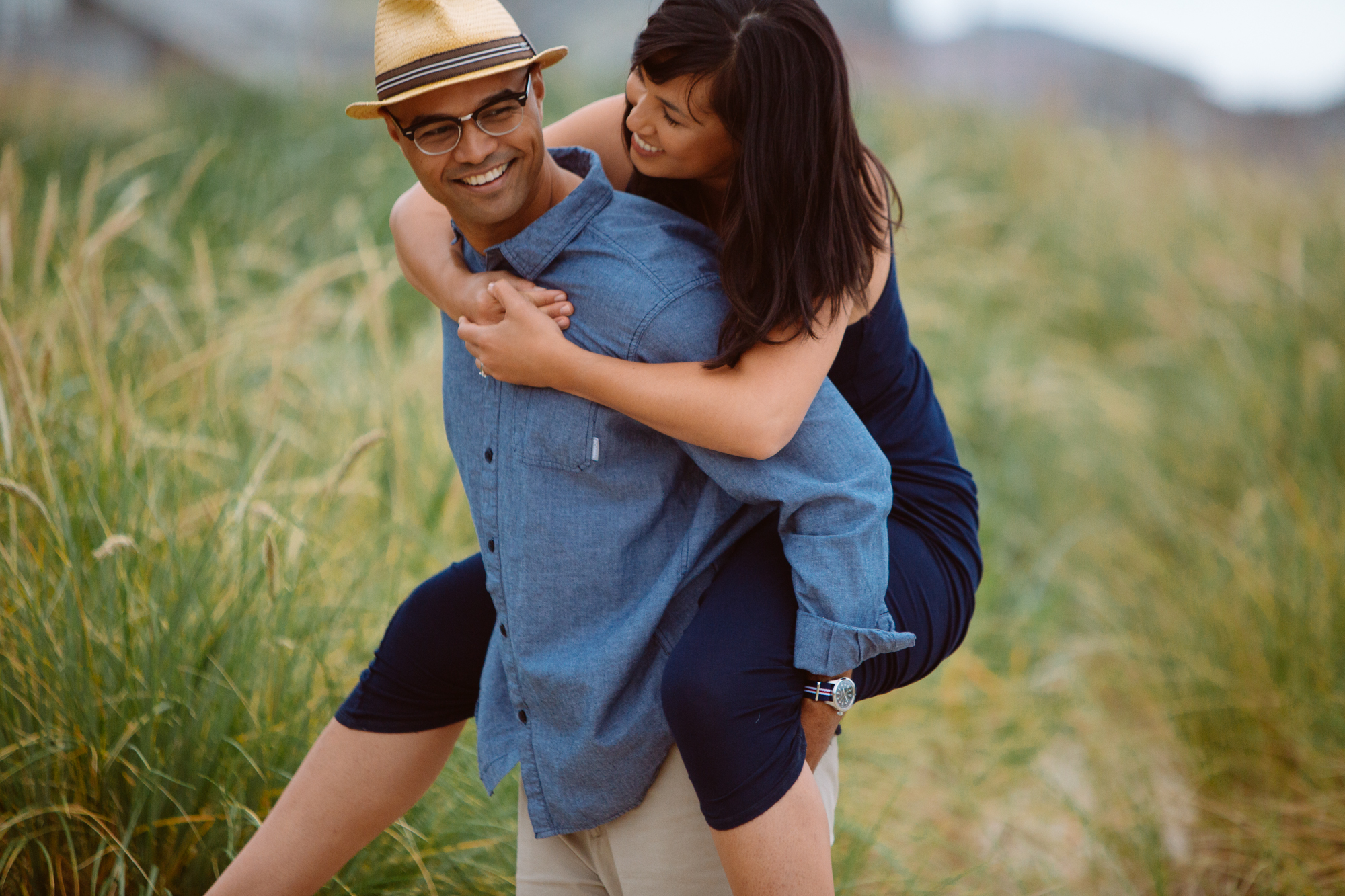 A woman in a dark dress is giving a piggyback ride to a man in a blue shirt and straw hat; they are smiling in a grassy outdoor setting. Jessica Hill Photography