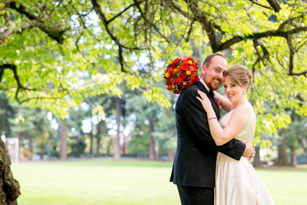 A couple in wedding attire poses closely together under green trees in a park, with the groom holding a red and yellow bouquet. Jessica Hill Photography - Wedding Photographer - Oregon