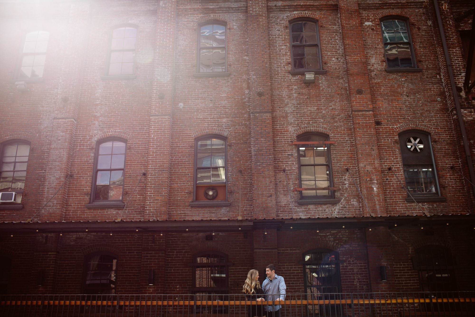 A couple stands on a balcony in front of a large brick building with tall windows on a sunny day. Jessica Hill Photography
