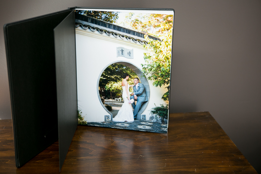 An open photo album on a wooden table displays a wedding photo of a bride and groom posing in a circular doorway with greenery in the background. Jessica Hill Photography - Wedding Photographer - Oregon