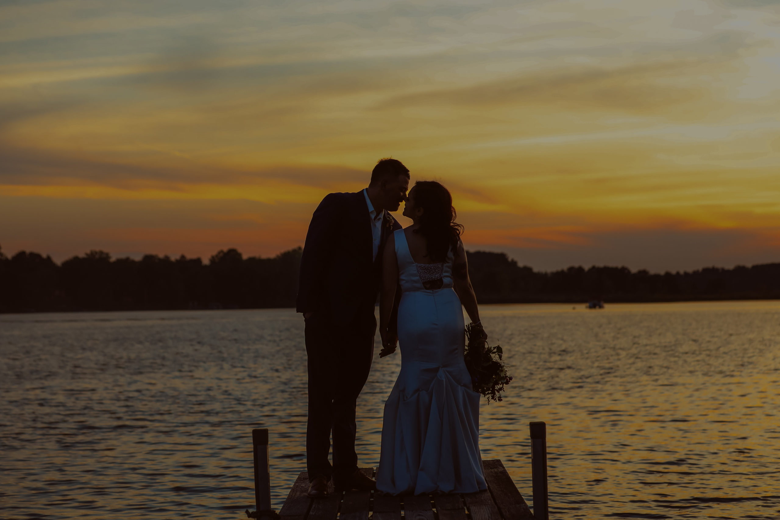 A couple in formal attire stands on a dock by a lake at sunset, holding hands and facing each other. Jessica Hill Photography