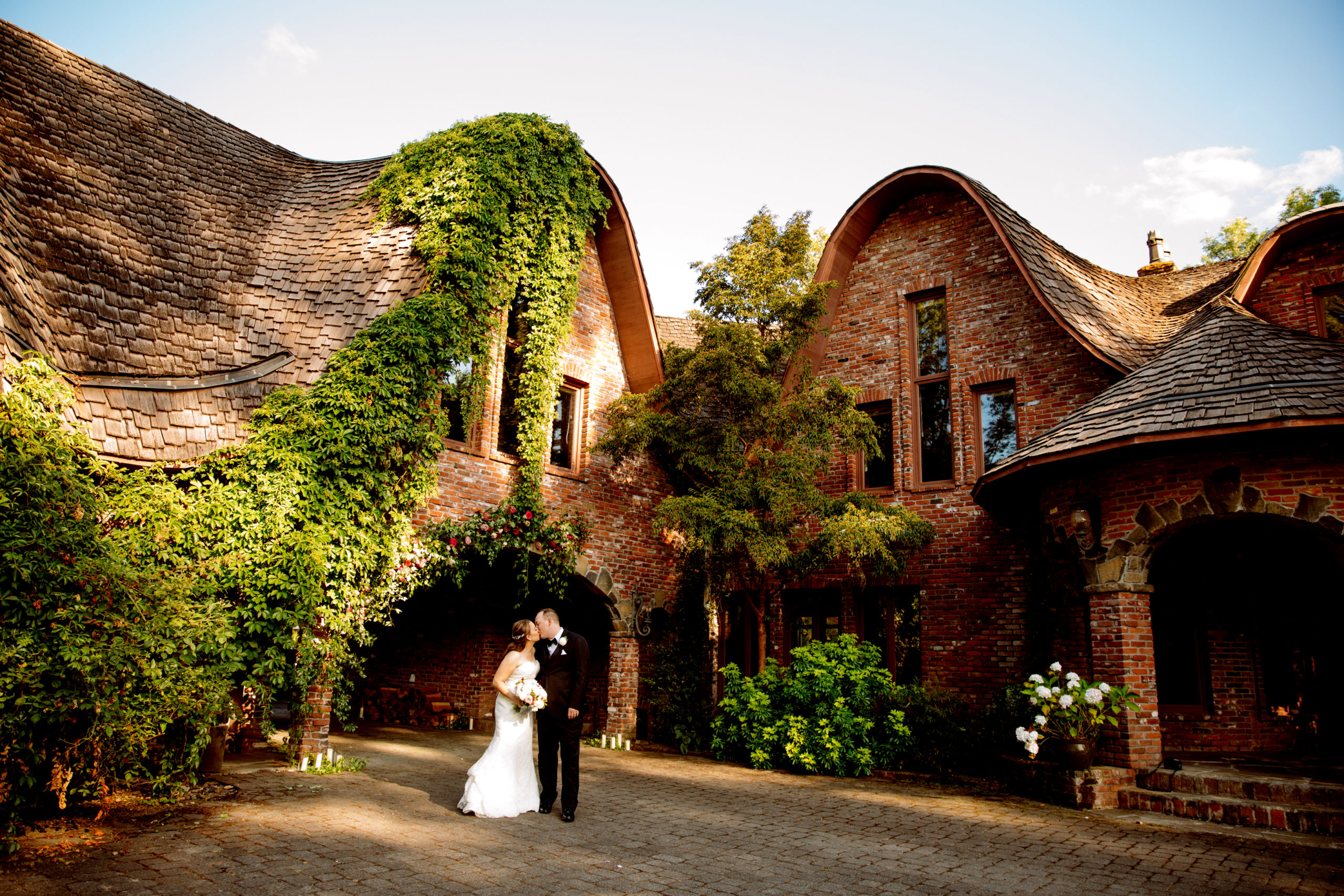 A bride and groom stand together in front of an ivy-covered brick building with arched roofs, surrounded by trees and soft evening light. Jessica Hill Photography