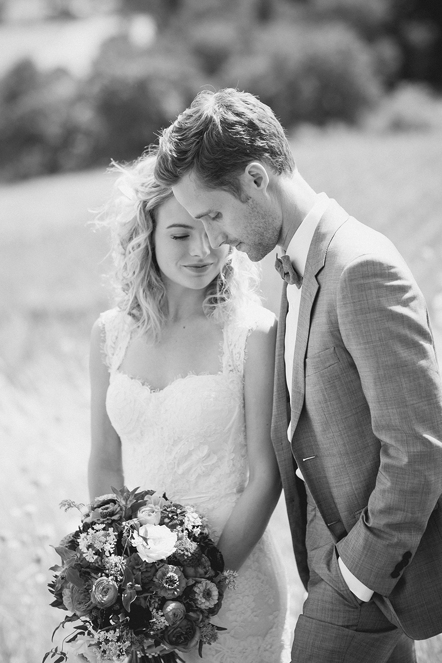 A bride in a lace wedding dress holding a bouquet stands next to a groom in a suit and bow tie. They gently lean their heads together outdoors, sharing an intimate moment. The photo is in black and white. Jessica Hill Photography - Wedding Photographer - Oregon