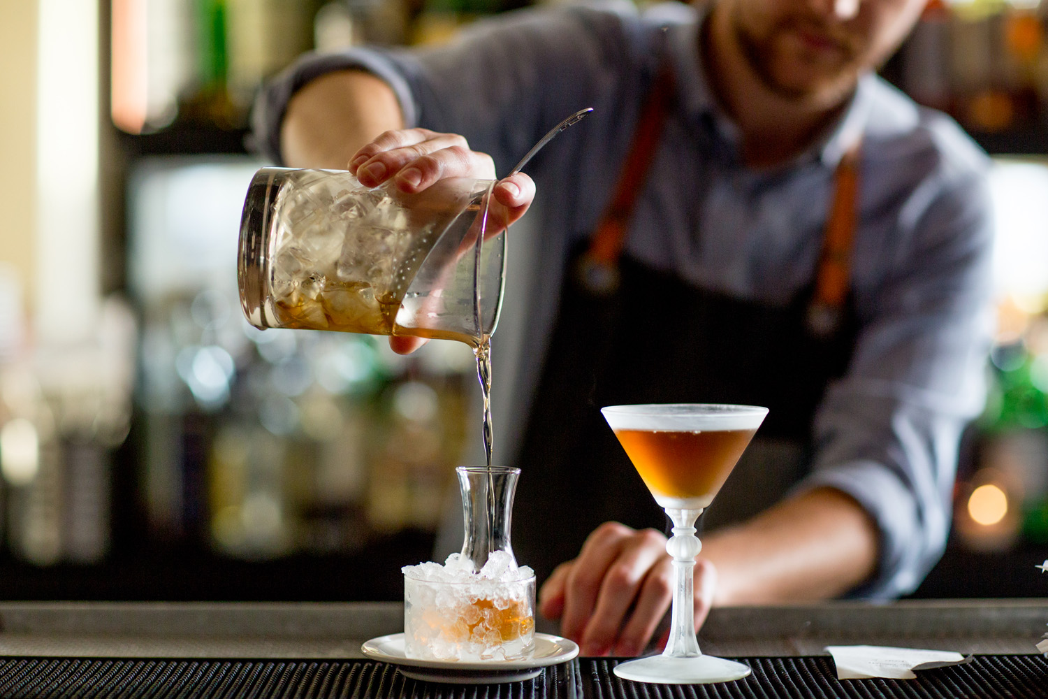 A bartender pours a cocktail from a mixing glass into a carafe filled with ice, next to a filled coupe glass on the bar. The bartender wears a gray shirt and a dark apron. Jessica Hill Photography - Wedding Photographer - Oregon