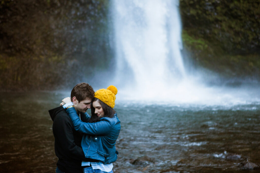 A couple embraces closely by a waterfall, with mist rising behind them. The woman wears a yellow knit hat and blue jacket, and the man wears a black hoodie. The scene is intimate and serene, set near the water's edge. Jessica Hill Photography - Wedding Photographer - Oregon