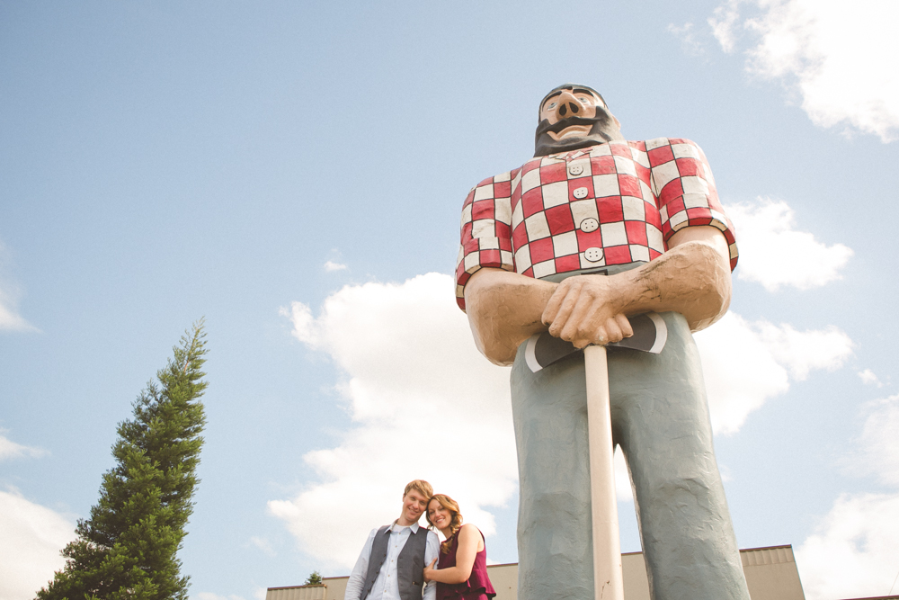 A couple stands smiling at the base of a large Paul Bunyan statue under a partly cloudy sky. Jessica Hill Photography - Wedding Photographer - Oregon