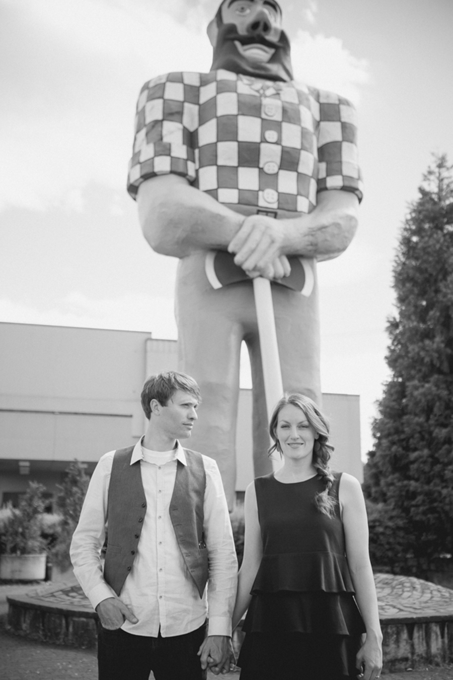 A man and woman hold hands in front of a large lumberjack statue, with buildings and trees visible in the background. The photo is in black and white. Jessica Hill Photography - Wedding Photographer - Oregon