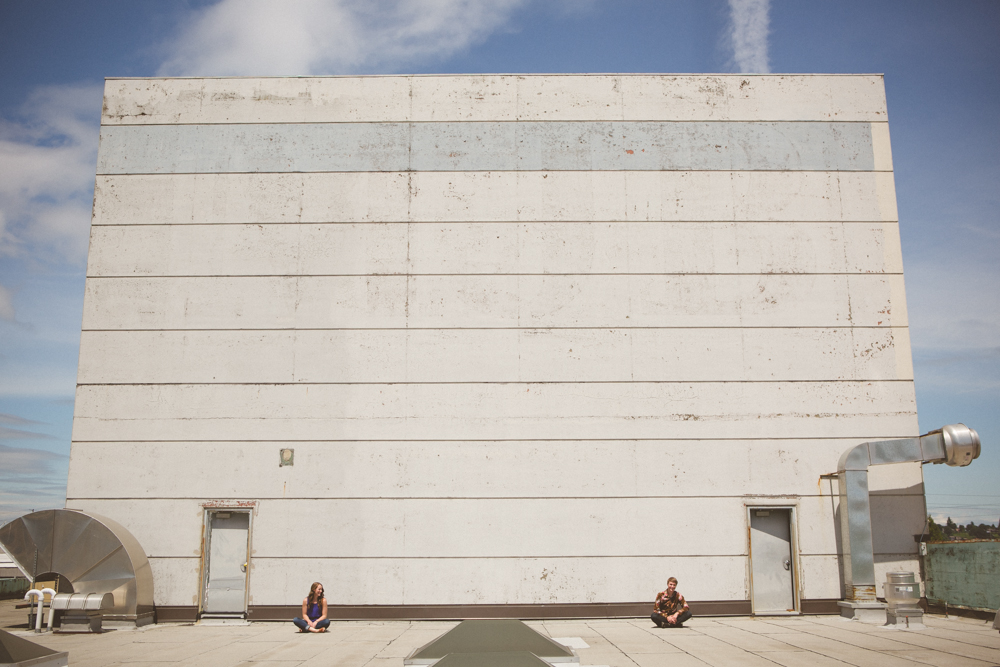 Two people are sitting far apart on a rooftop in front of a large, plain, weathered white wall with two doors and industrial vents. Jessica Hill Photography - Wedding Photographer - Oregon