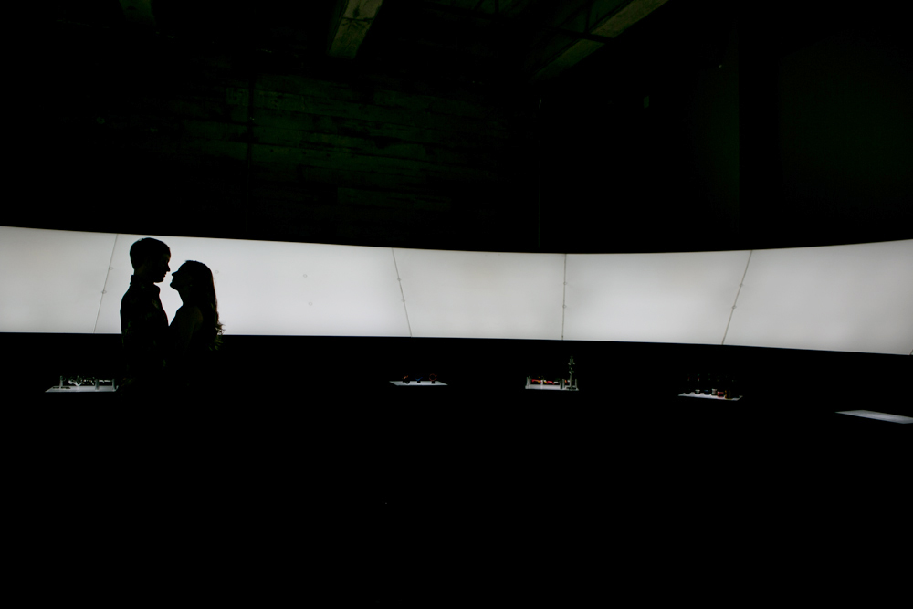 Two people stand in silhouette facing each other in front of a large, curved, brightly lit screen in an otherwise dark room. Jessica Hill Photography - Wedding Photographer - Oregon