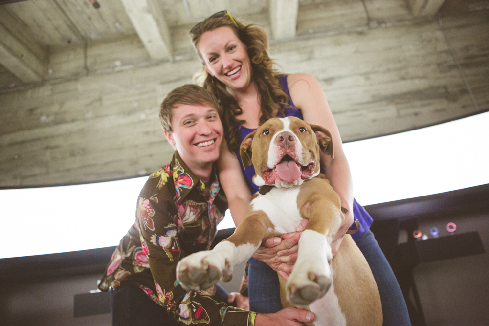 Two people smiling while holding a large brown and white dog towards the camera in an indoor setting with a concrete ceiling. Jessica Hill Photography - Wedding Photographer - Oregon