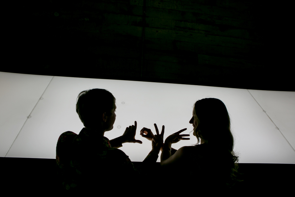 Two people are silhouetted against a bright rectangular light, using their hands to form shapes or letters in front of the illuminated background. Jessica Hill Photography - Wedding Photographer - Oregon