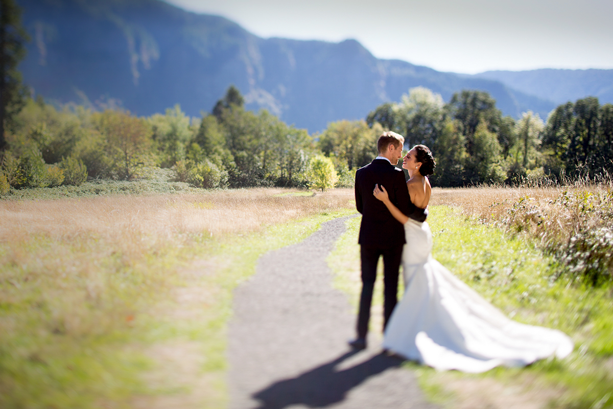 A bride and groom stand on a path in a sunlit field, embracing and looking at each other, with trees and mountains in the background. The bride wears a white dress; the groom is in a dark suit. Jessica Hill Photography - Wedding Photographer - Oregon