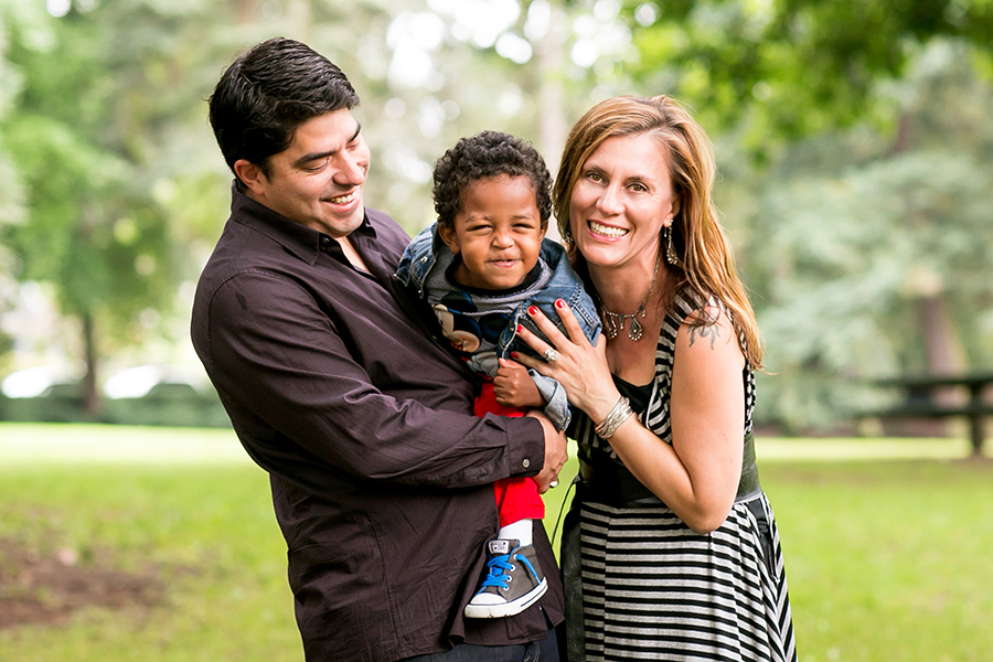 A man and woman stand outdoors, smiling and holding a young child between them. The family appears happy, with greenery and blurred trees in the background on a bright day. Jessica Hill Photography - Wedding Photographer - Oregon
