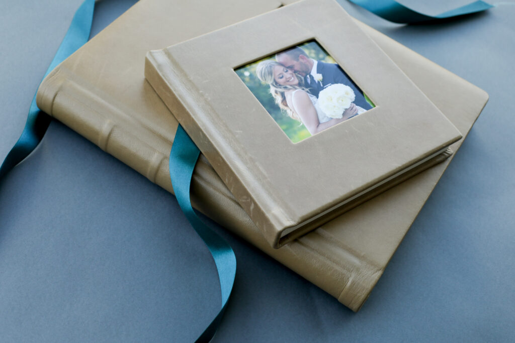 Two beige photo albums with teal ribbons, one featuring a cover photo of a bride and groom, are stacked on a gray surface. Jessica Hill Photography