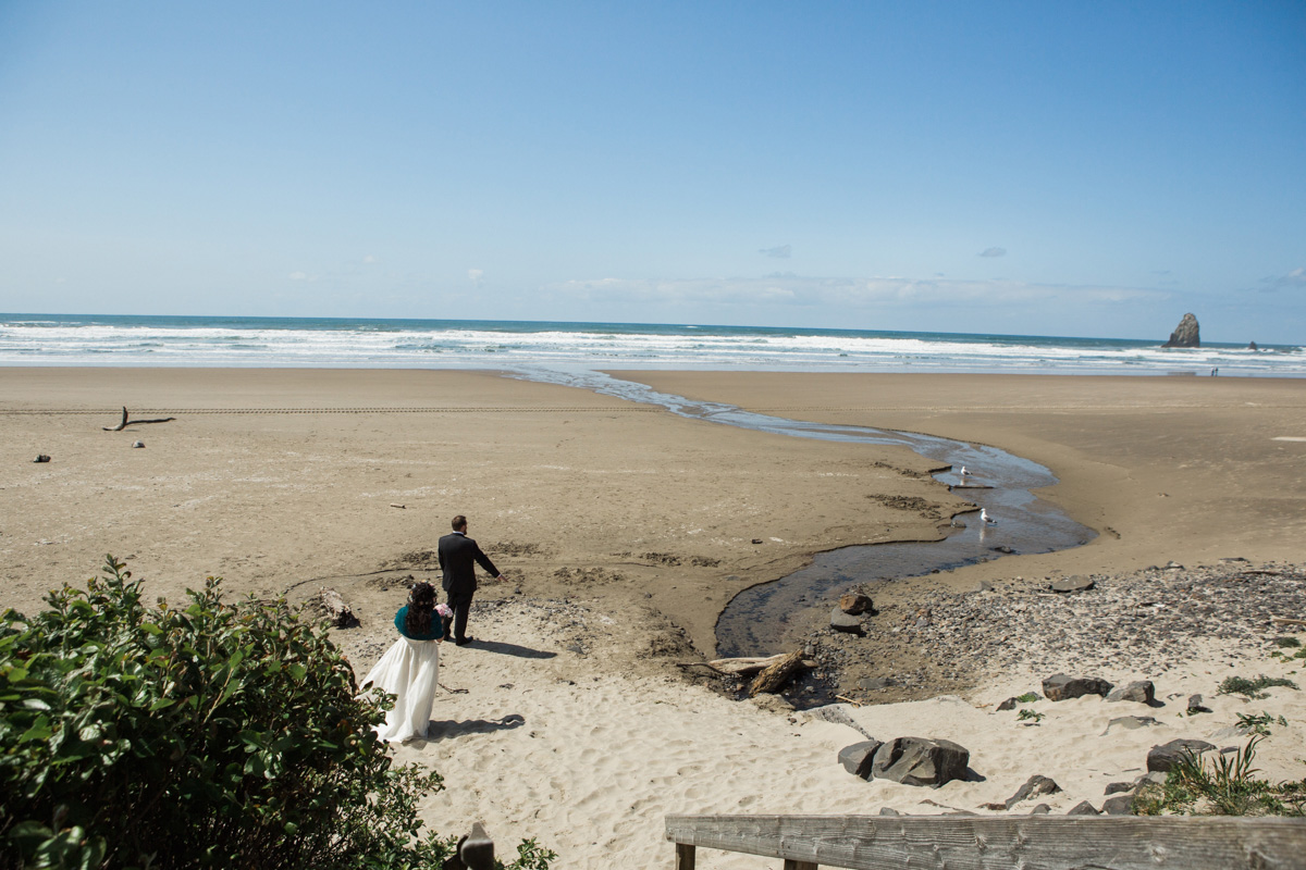 A bride and groom walk hand in hand on a sandy beach toward the ocean under a clear blue sky, with scattered driftwood and a small stream flowing to the sea. Jessica Hill Photography - Wedding Photographer - Oregon
