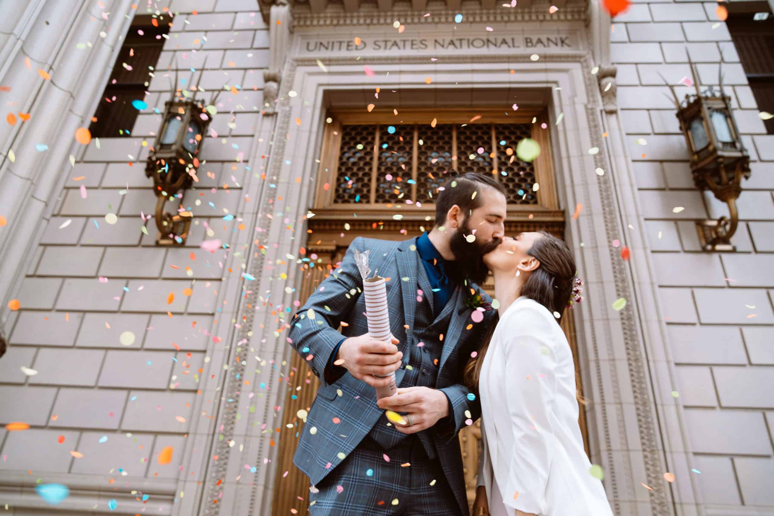 Couple kissing amidst colorful confetti at bank entrance.