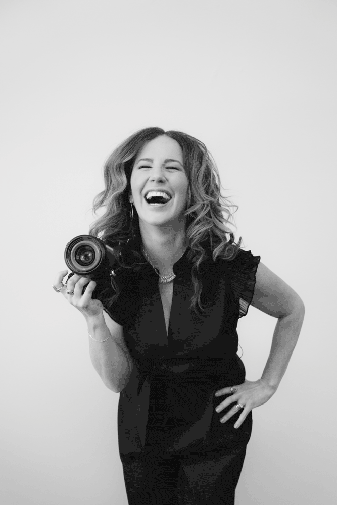 Woman with wavy hair wearing a dark blouse smiles and laughs while holding a camera in her right hand, standing against a plain white background—capturing the joyful spirit of Jessica Hill Photography. Jessica Hill Photography
